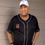 Man wearing a black baseball jersey with red lettering and a white cap, standing against a beige wall.