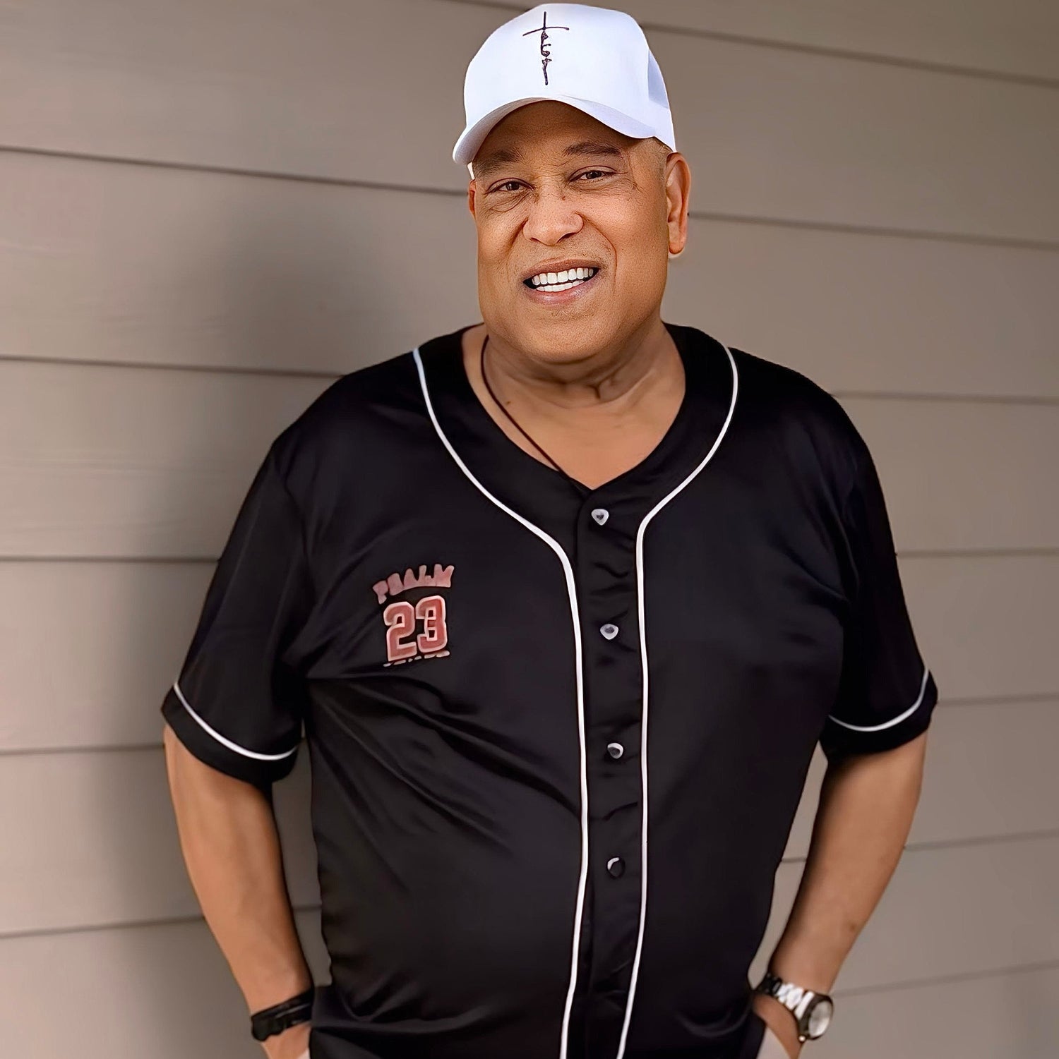 Man wearing a black baseball jersey with red lettering and a white cap, standing against a beige wall.