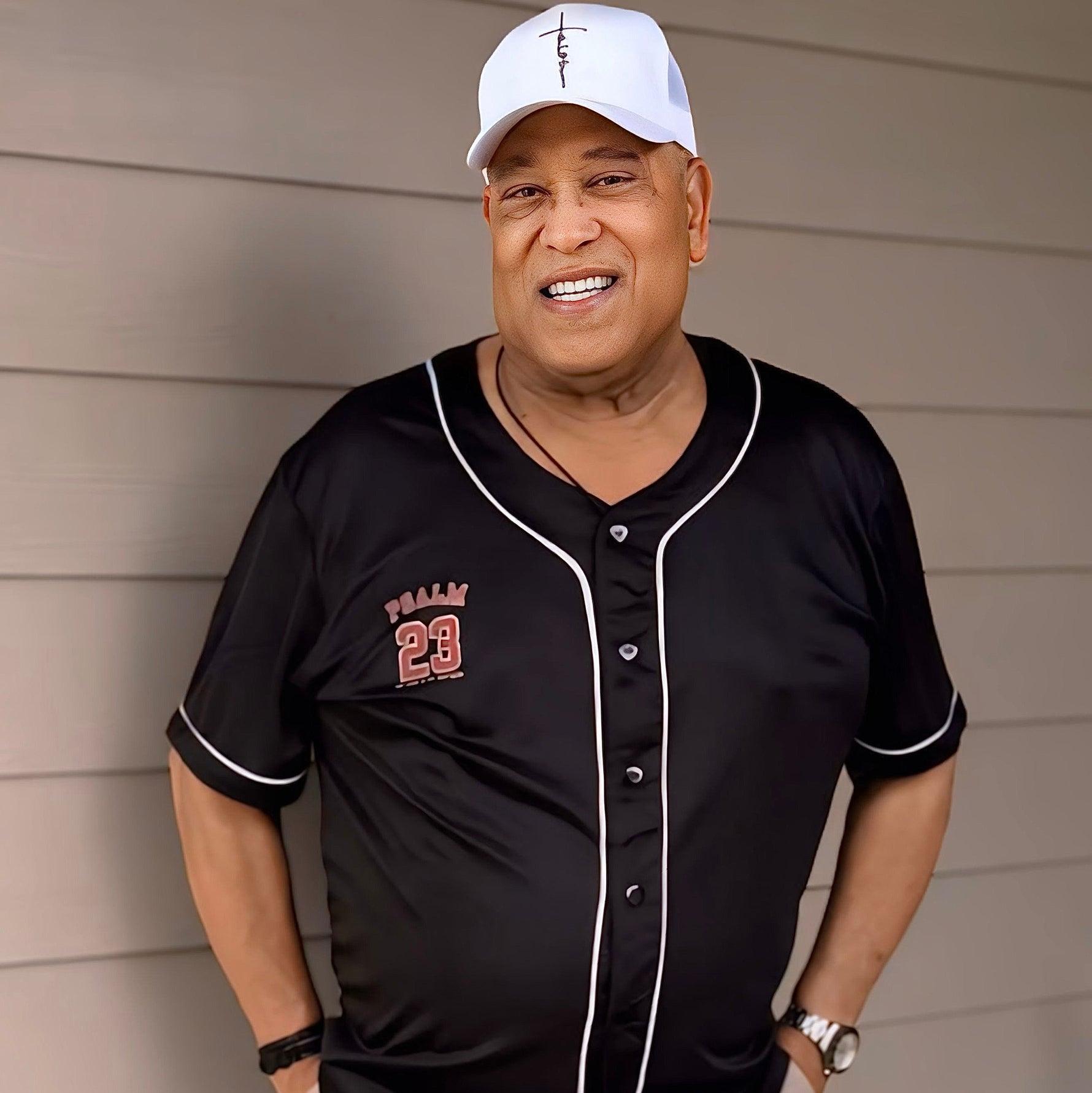 Man wearing a black baseball jersey with red lettering and a white cap, standing against a beige wall.