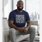 Man wearing a navy blue t-shirt with 'Purpose Over Popularity' text, sitting in a modern living room.