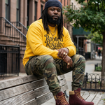 Person wearing a yellow 'Rooted in Royalty' sweatshirt sitting on a bench in an urban setting.