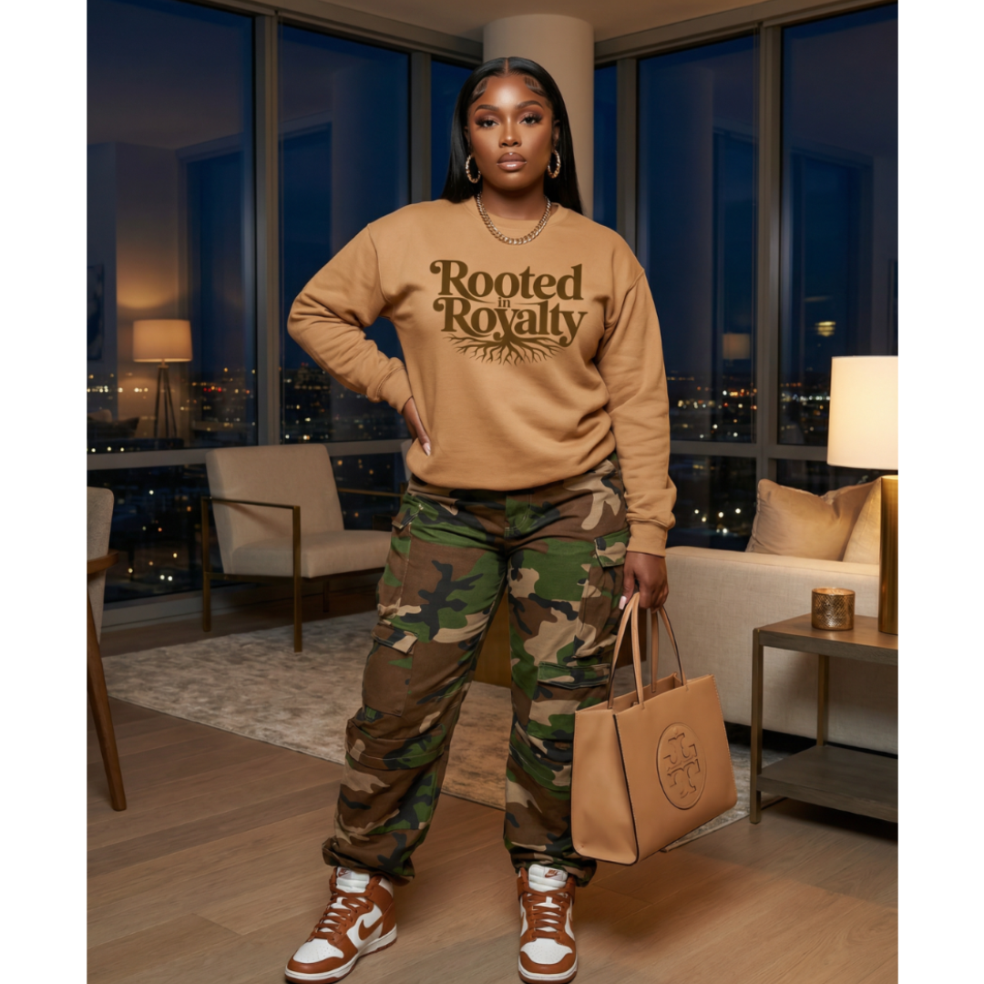 Person wearing a beige sweatshirt with text and camouflage pants, holding a brown bag in a room with city lights.
