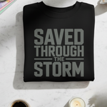 Black sweatshirt with 'Saved Through The Storm' text on a light surface with coffee and books.