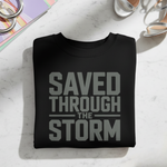 Black sweatshirt with 'Saved Through The Storm' text on a light surface with coffee and books.