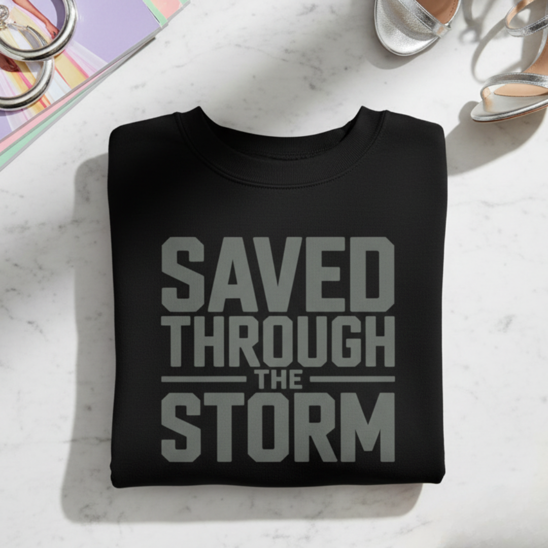 Black sweatshirt with 'Saved Through The Storm' text on a light surface with coffee and books.