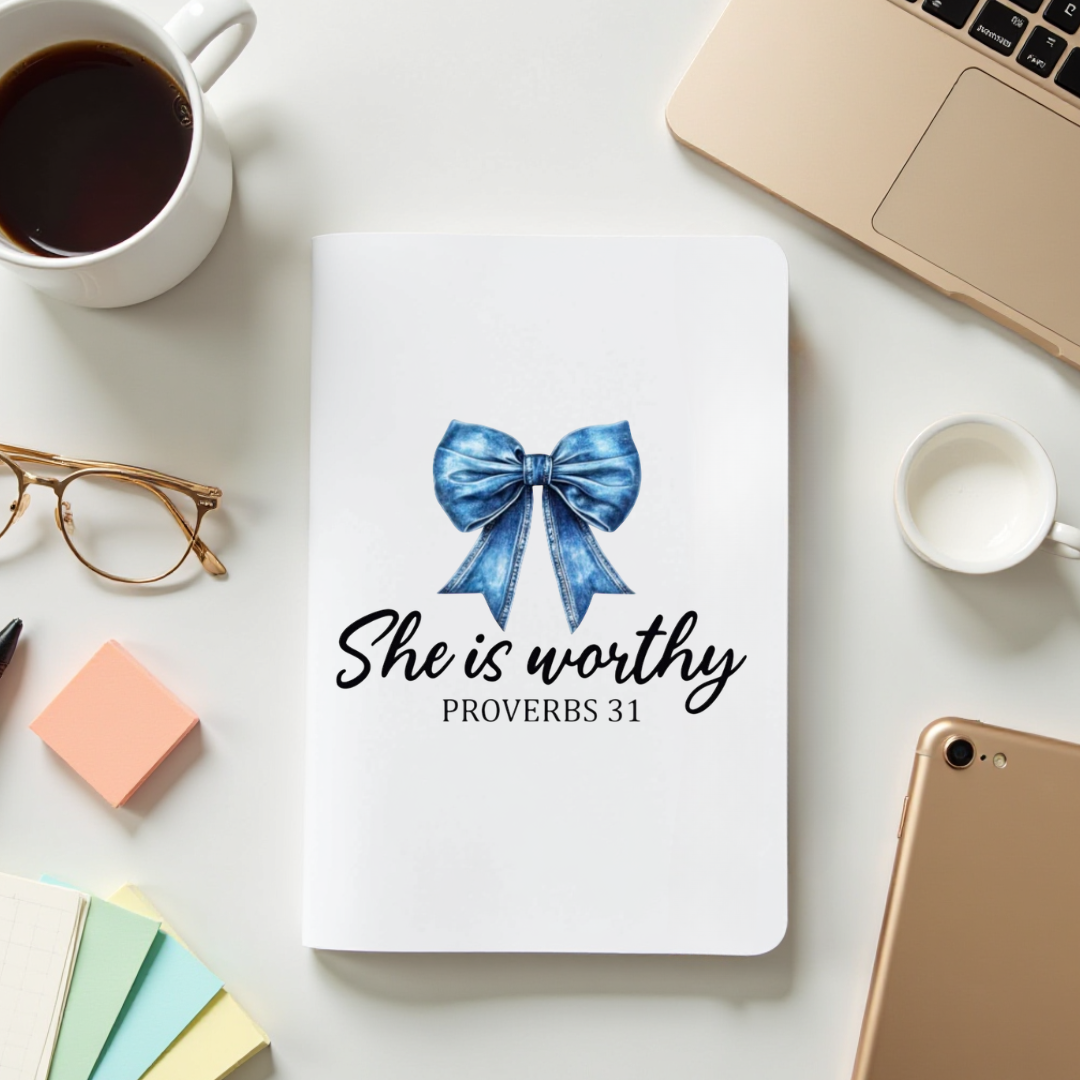 Notebook with a blue bow illustration and 'She is worthy Proverbs 31' text on a desk with coffee, glasses, and phone.
