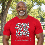 A smiling man wearing a bright red graphic t-shirt with 'SON OF THE KING' in metallic silver lettering and a Bible verse printed below, set against a blurred outdoor park background.