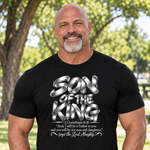 A smiling man wearing a black graphic t-shirt with 'SON OF THE KING' and a Bible verse printed in silver and white, set against a blurred outdoor background of trees and grass.