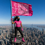 A woman stands on a high-rise balcony overlooking a city skyline, holding a bright pink flag with white text. She is wearing a matching bright pink T-shirt, black pants, and pink boots, with a black face mask.