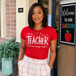 A woman wearing a red graphic t-shirt that says 'TEACHER teach encouragement inspire' with a white plaid skirt, standing in front of a school classroom door.