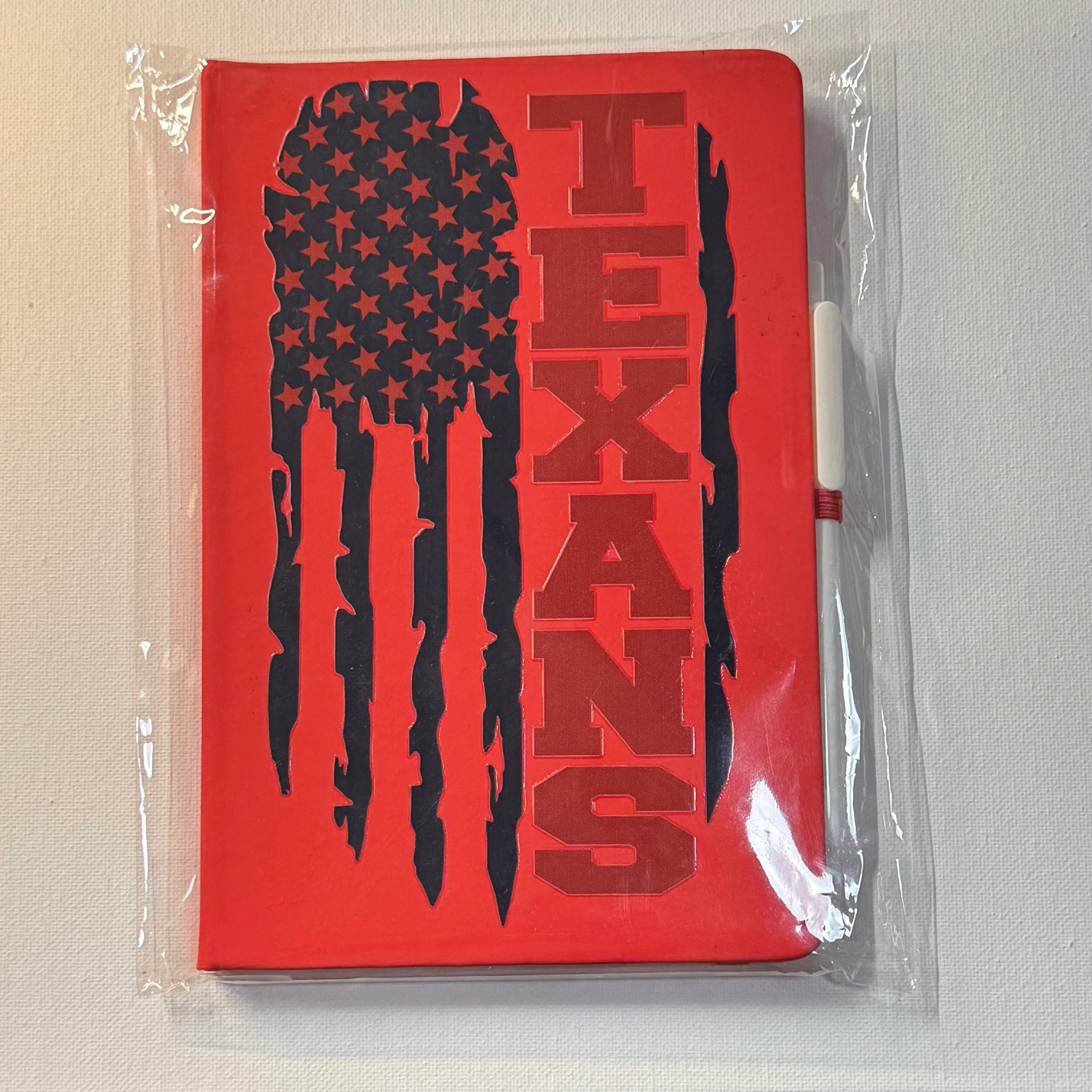 Red notebook with 'TEXANS' and American flag design in clear packaging on a white surface.