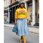 Woman wearing a yellow hoodie with text, blue skirt, and yellow boots on a city street.