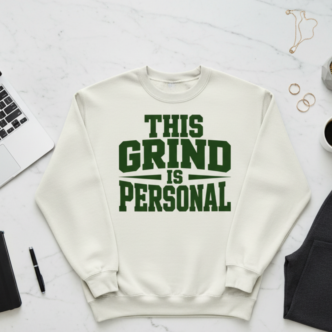 White sweatshirt with 'This Grind is Personal' text on a light gray surface with office items.