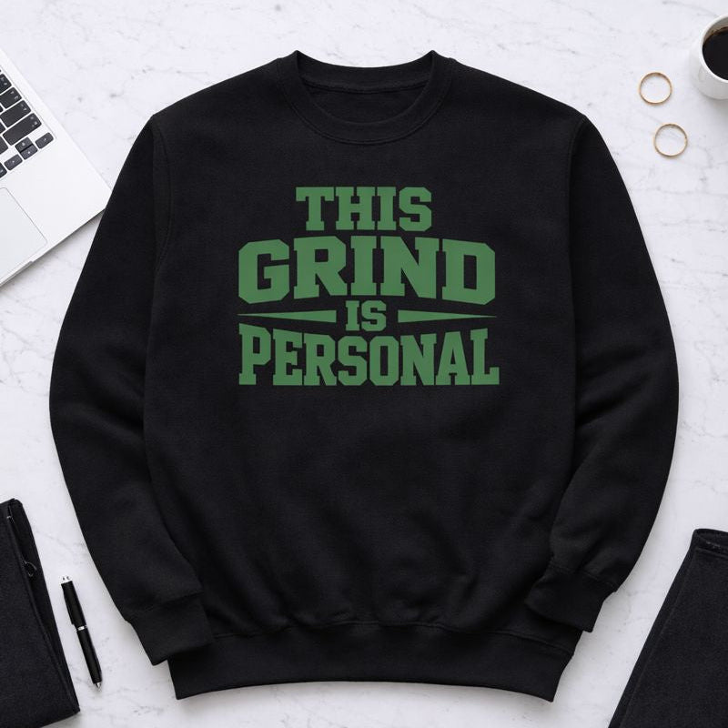Black sweatshirt with 'This Grind is Personal' text on a white surface with office supplies.