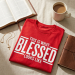 Folded red t-shirt with 'This is what blessed looks like' text on a table with a book and coffee cup.