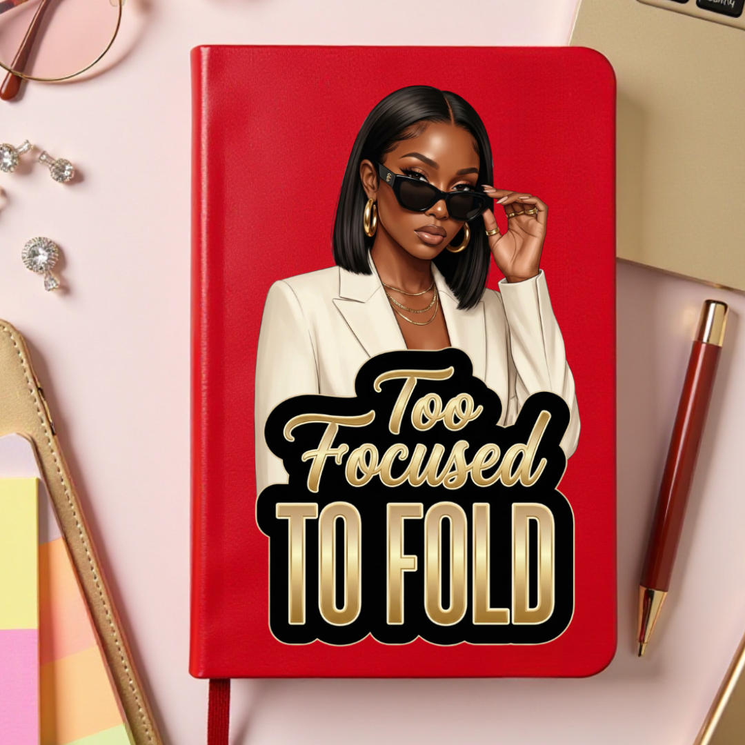 Red notebook with a graphic of a woman and 'Too Focused To Fold' text, surrounded by office supplies on a light pink background.