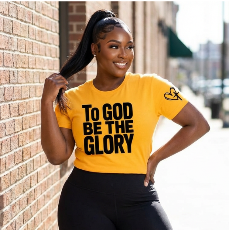 Woman wearing a yellow t-shirt with 'To God Be the Glory' text, standing against a brick wall.