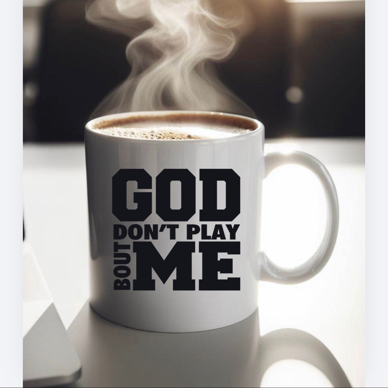 White mug with 'GOD DON'T PLAY ABOUT ME' text, steaming coffee inside, on a blurred background.