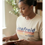 Woman wearing a 'Unafraid' t-shirt, sitting at a desk with books and a plant in the background.