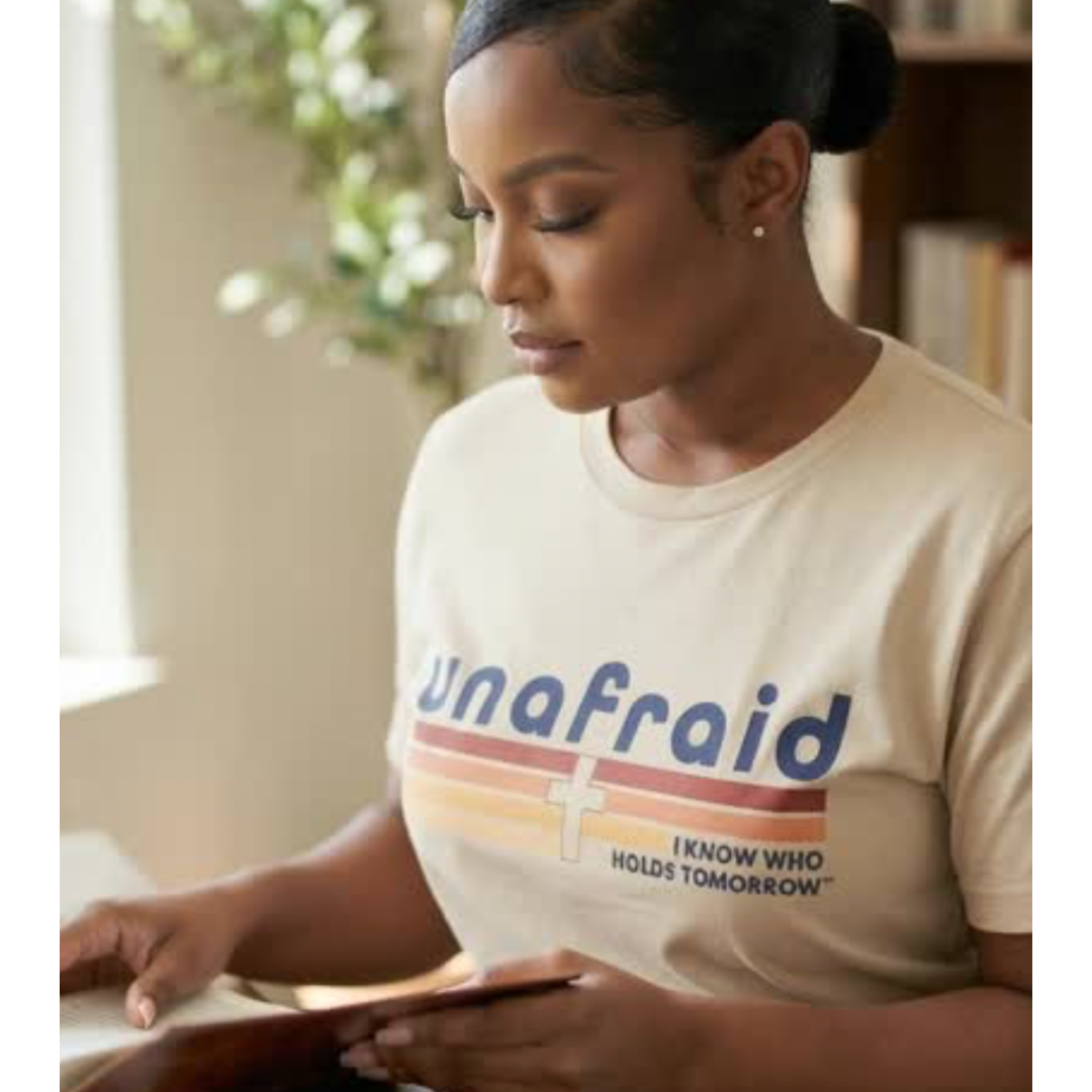 Woman wearing a 'Unafraid' t-shirt, sitting at a desk with books and a plant in the background.