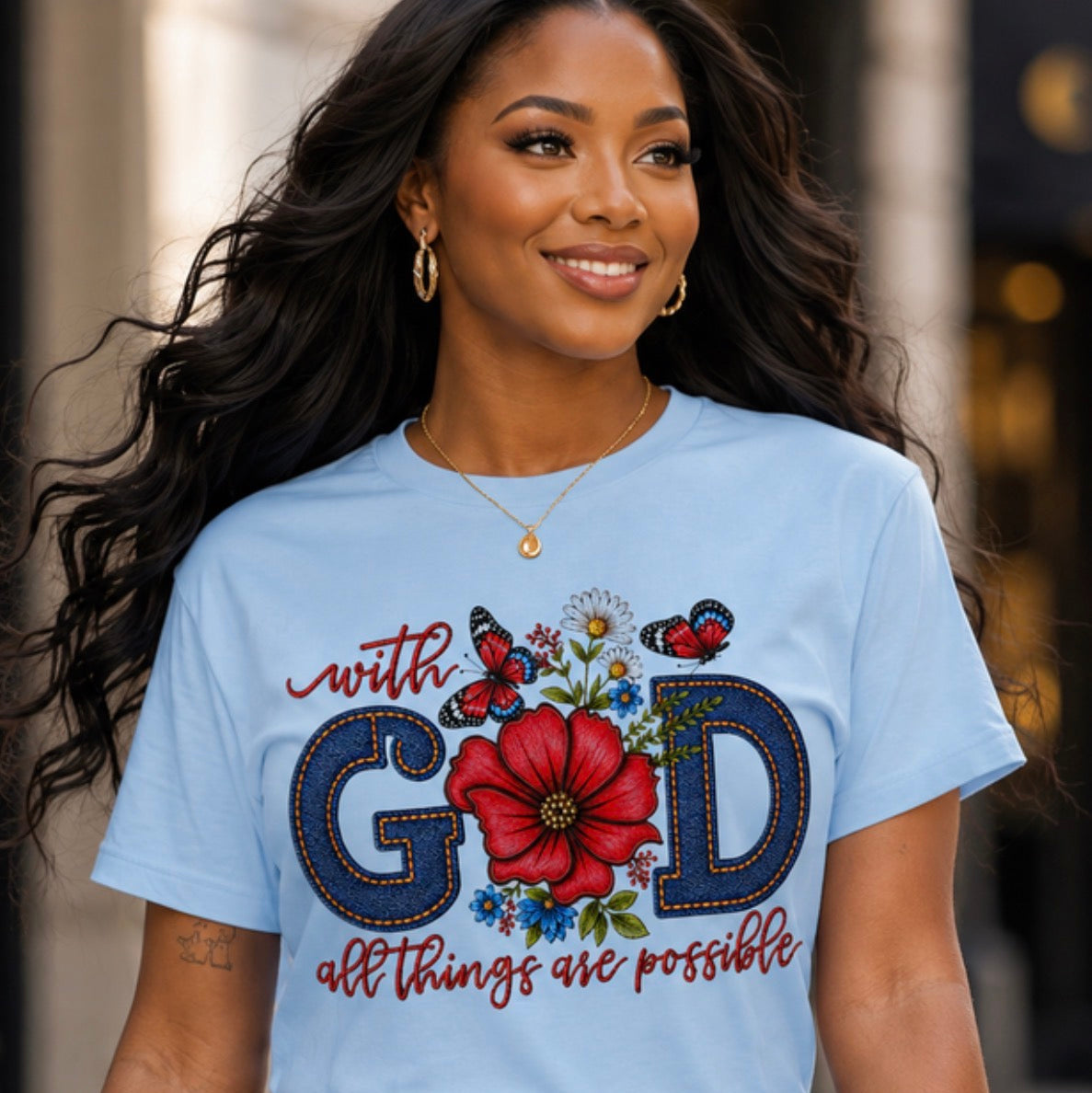 A woman wearing a light blue graphic t-shirt with a colorful 'with GOD all things are possible' design, paired with blue jeans in an outdoor urban setting.