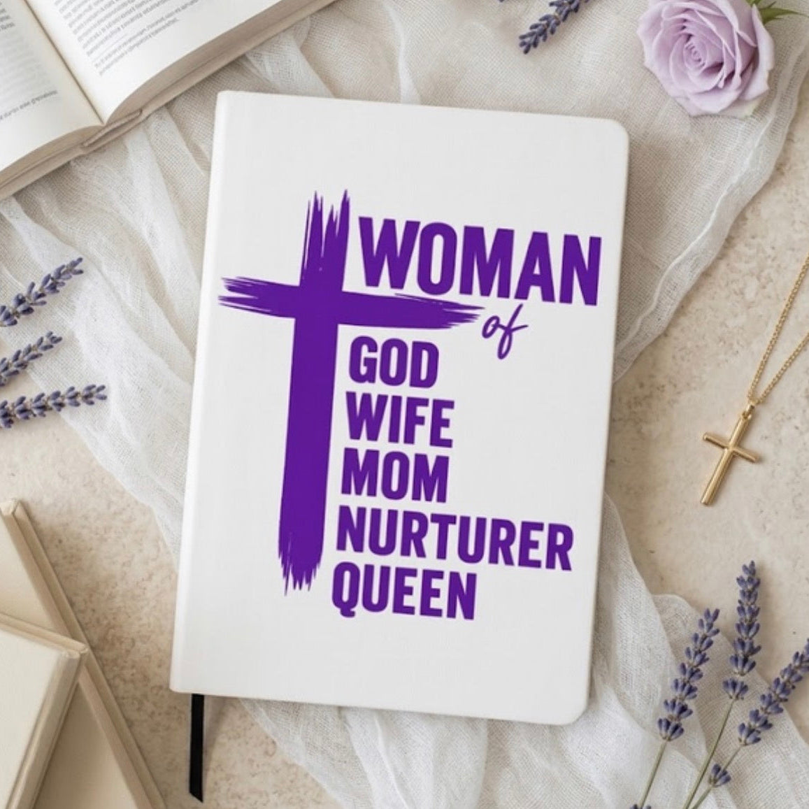 A white hardcover journal with purple text reading 'WOMAN of GOD WIFE MOM NURTERER QUEEN' and a large purple cross, styled on a beige fabric surface with lavender sprigs, an open book, a pink rose, and a gold cross necklace.