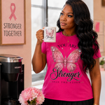 A woman in a bright pink graphic t-shirt with a butterfly design and the words 'YOU ARE Stronger THAN THE STORM' holds a white mug with a matching pink butterfly graphic. She is standing in a kitchen-like setting with a Keurig coffee maker, a framed 'Stronger Together' sign, and pink flowers on a table.