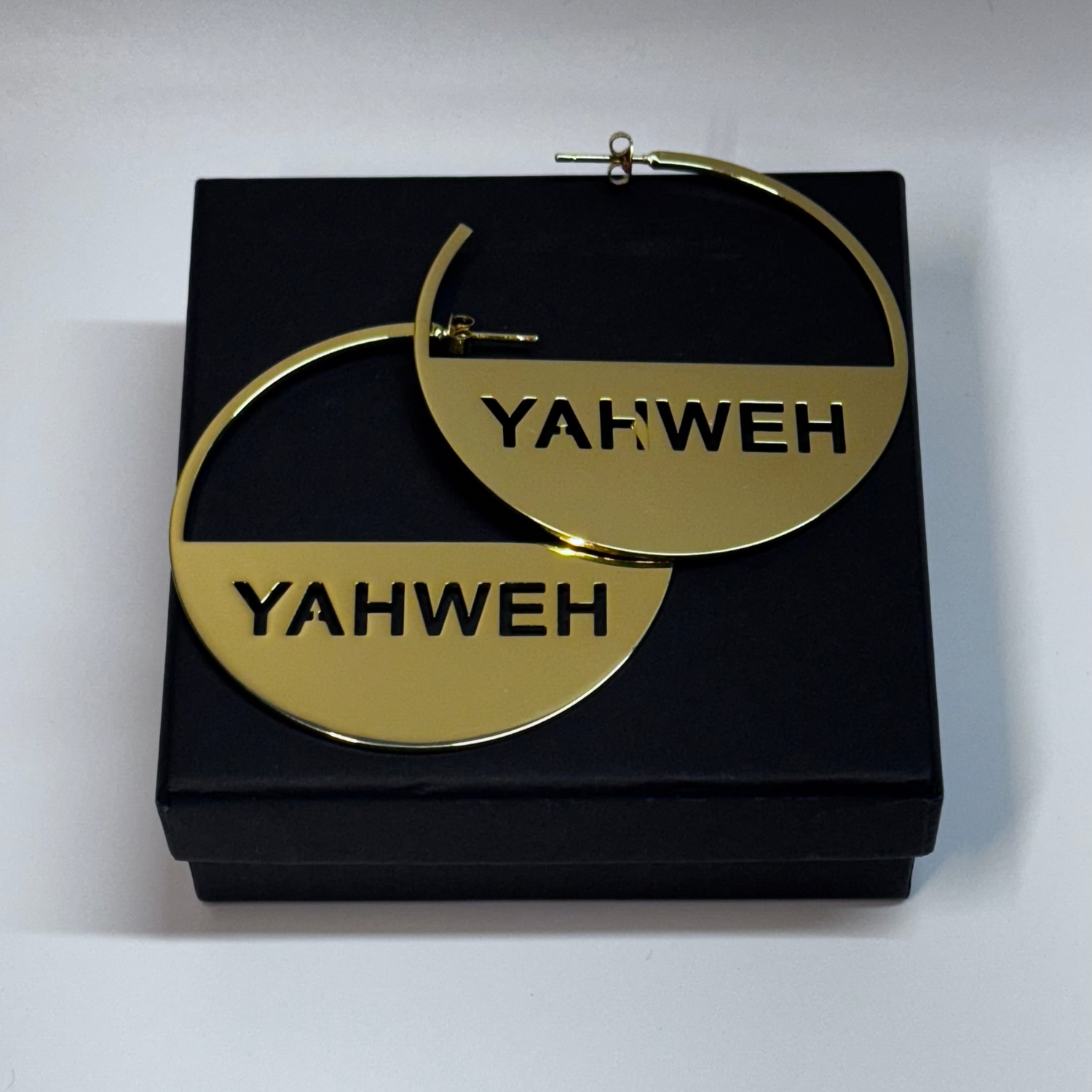 Gold hoop earrings with 'YAHWEH' engraving on a black box against a white background