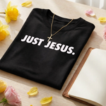 Black t-shirt with 'JUST JESUS.' text on a wooden surface with flowers and an open book.