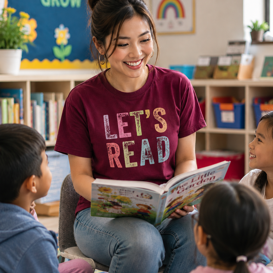 A smiling woman in a maroon graphic T-shirt reading to two children in a bright classroom. The shirt has the words 'LET'S READ' in large multicolored letters.