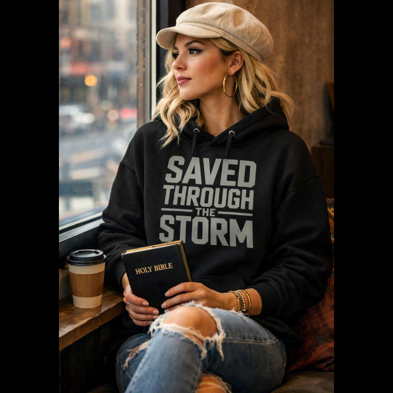 Person wearing a hoodie with 'Saved Through the Storm' text, holding a Bible, sitting by a window.