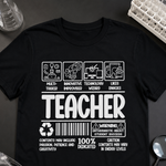 A black short-sleeve graphic T-shirt laid flat on a dark surface, surrounded by small study and science props like glasses, a magnifying glass, and molecular model beads. The shirt has large white text reading 'TEACHER' with humorous and educational icons above and below, plus a barcode and warning-style print.