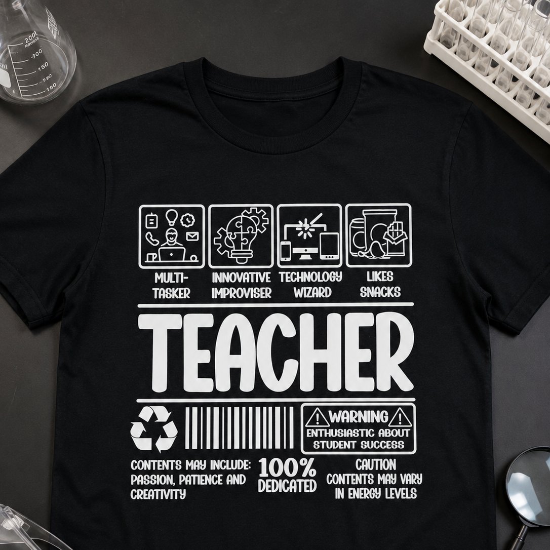 A black short-sleeve graphic T-shirt laid flat on a dark surface, surrounded by small study and science props like glasses, a magnifying glass, and molecular model beads. The shirt has large white text reading 'TEACHER' with humorous and educational icons above and below, plus a barcode and warning-style print.