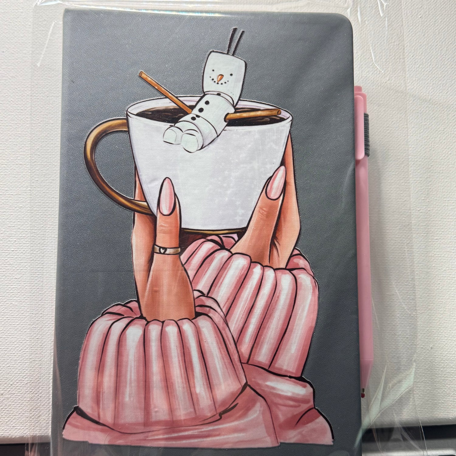 Notebook with a design of hands holding a mug with marshmallows, on a plain background.